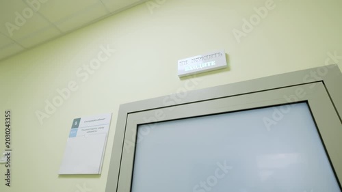 Modern clinic corridor displays digital cue panel with Cyrillic text over office entrance. Bright ceiling lights create pristine atmosphere