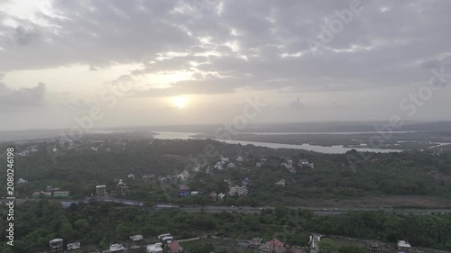 Aerial view of houses in Old Goa surrounded by lush greenery during the pre-monsoon season, with the Mandovi River in the background and a dramatic sunset beneath cloudy skies.