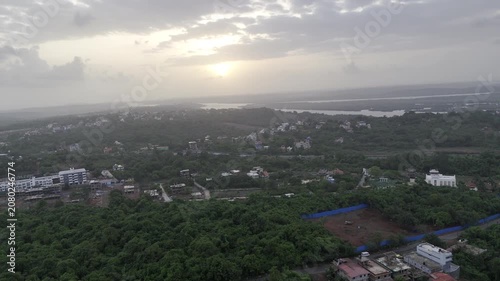 Aerial view of houses in Old Goa surrounded by lush greenery during the pre-monsoon season, with the Mandovi River in the background and a dramatic sunset beneath cloudy skies.