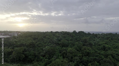 Aerial view of houses in Old Goa surrounded by lush greenery during the pre-monsoon season, with the Mandovi River in the background and a dramatic sunset beneath cloudy skies.