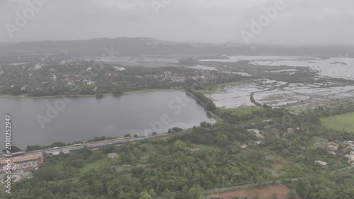 Aerial view of a train at Karmali Railway Station near Carambolim Lake in Old Goa, surrounded by lush greenery, coconut palms, and trees beneath cloudy monsoon evening skies.