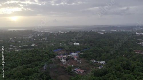 Aerial view of houses in Old Goa surrounded by lush greenery during the pre-monsoon season, with a dramatic sunset glowing through cloudy skies over the scenic landscape.
