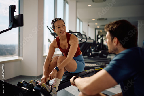 Tableau sur toile Happy Korean sportswoman talking to her personal trainer while working out on rowing machine in gym