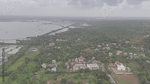 Aerial view of Old Goa and the Mandovi River during the monsoon season, featuring coconut palms, green fields, backwaters, lush greenery, nearby houses, and cloudy skies.