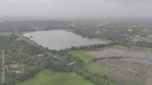 Aerial view of a train at Karmali Railway Station near Carambolim Lake in Old Goa, surrounded by lush greenery, coconut palms, and trees beneath cloudy monsoon evening skies.