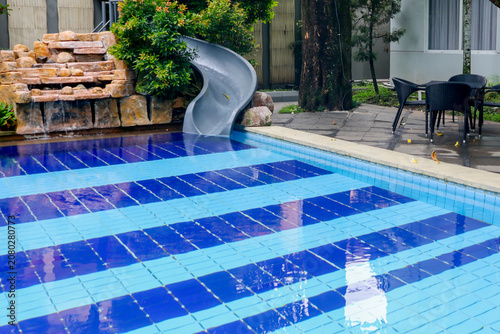 Blue tiled pool gleams under sunlight. Curved slide invites joy and play. Stone waterfall adds natural charm. Greenery frames the tranquil scene. Outdoor seating offers relaxing pause