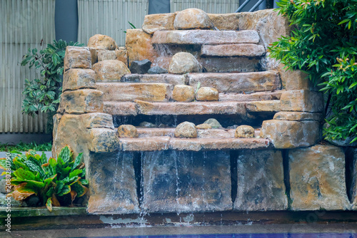 Stone waterfall cascades gently over tiered rock formations. Green foliage frames the natural water feature. Water pools softly at the base, reflecting calm surroundings