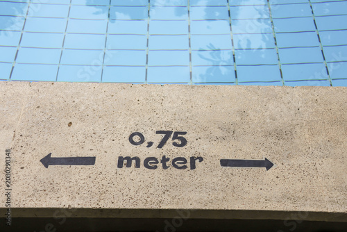 Clear blue pool tiles reflect sky above. Concrete edge shows 0.75 meter marker with arrows. Pool depth guide helps swimmers measure distance. Ideal for swimming facility or training center