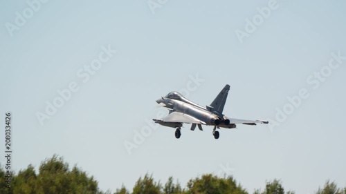 Combat aircraft on final approach with gear down and landing lights on in slow motion, military fighter jet seen head on against blue sky, warplane coming in to land