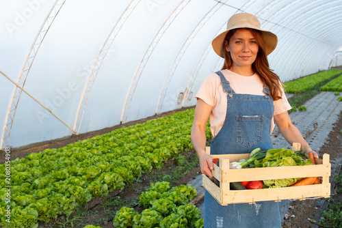 Woman farmer in straw hat holding crate of fresh vegetables inside greenhouse
