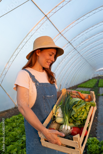 Smiling farmer in straw hat holding a crate full of fresh vegetables inside a greenhouse