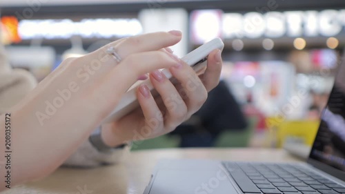 Person uses smartphone in public space. Fingers tap screen with focus. Laptop sits nearby on table. Background shows busy indoor setting. Scene captures modern digital multitasking