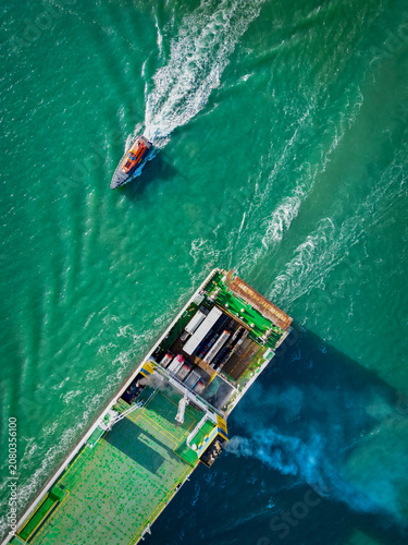 Aerial view of a large cargo ship navigating Dublin Harbour with a pilot boat nearby, leaving white wake trails in the green water of Dublin, County Dublin, Ireland.