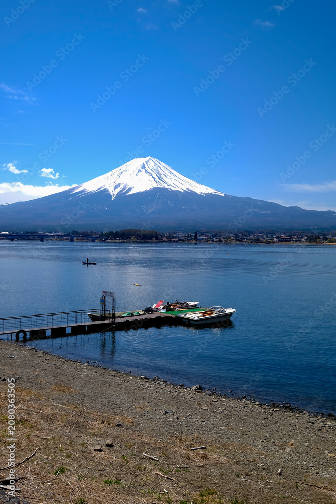 Fototapeta premium Snowy Fuji mountain lake pier boat blue sky sakura shoreline village landscape Snowy Fuji mountain above calm lake with pier and boat near shoreline village under blue sky with sakura season scenery