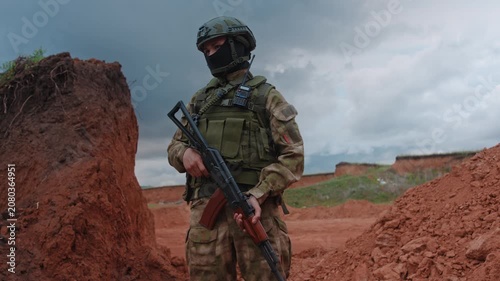 Uniformed soldier standing with rifle in low ready posture on red soil ridge beneath overcast sky, side view showing helmet, camouflage uniform, alert focused stance