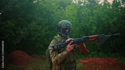 Camouflaged soldier adjusting rifle in wooded clearing with tactical vest, helmet, goggles and radio, alert stance during military training in green brown forest environment