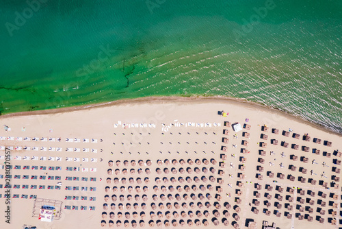 Aerial top down view of an organized sandy beach resort with rows of straw umbrellas and white sun loungers near the sea. Aerial top down view of rows of straw umbrellas and sun loungers on a sandy be