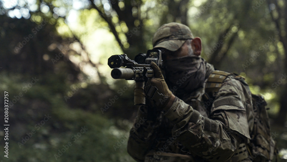Fototapeta premium Camouflaged soldier in a dark forest on a recon mission. Hiding and aiming with a AR-15 carbine rifle with a laser collimator and a magnifier