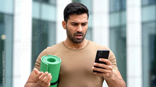 Confused young man holding a yoga mat and looking at his smartphone with a puzzled expression outdoors.