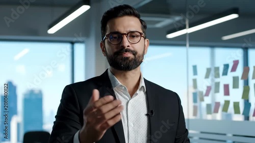 Confident businessman in glasses talking to camera in modern office setting.