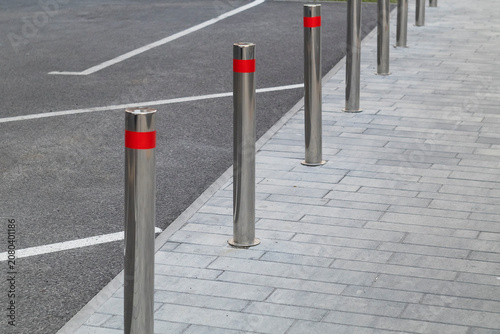 Row of modern stainless steel bollards with bright red reflective bands installed along a paved sidewalk to enhance pedestrian safety and restrict vehicle access.