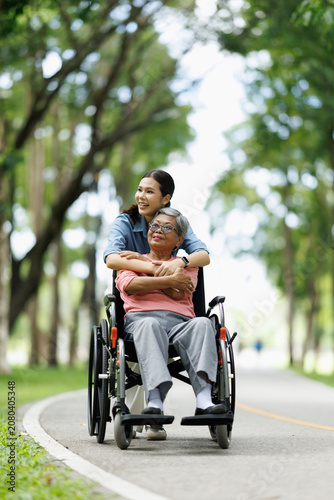 Asian daughter embracing her elderly mother who is sitting in a wheelchair, sharing a tender moment of care and affection during a leisurely outing in a lush green park