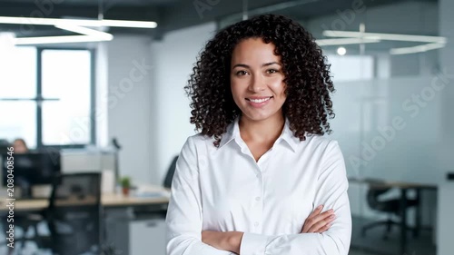 Smiling young businesswoman with crossed arms standing in modern office environment, looking at camera.