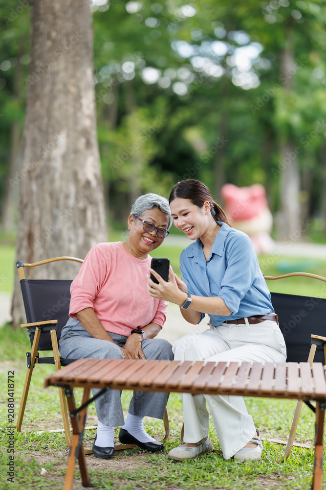 Fototapeta premium Granddaughter and grandmother sitting outdoors in a park, enjoying quality time together and sharing a happy moment while looking at a smartphone, bridging generations with technology