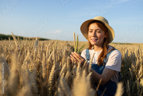 Woman farmer in straw hat inspecting ripe wheat field at sunset