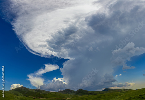 Tableau sur toile A large cumulonimbus cloud with a distinct anvil shape towering over the green, rolling hills