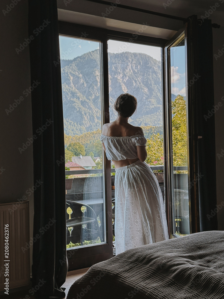 Fototapeta premium Woman in White Dress Gazing Out Window at Mountains