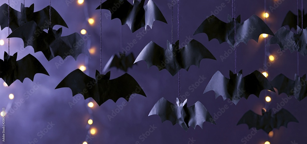 Fototapeta premium Against a rich purple background, black paper bats dangle amidst gentle glowing lights for Halloween decor.