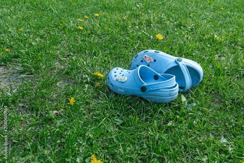 Blue rubber clogs with cute decorative charms layout on lush green grass field during bright summer day, high angle shot of casual kids footwear on lawn suitable for retail advertising