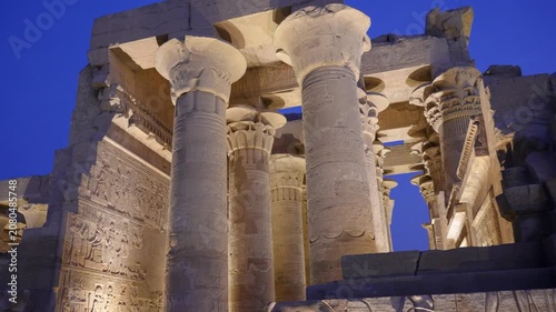 A low angle view of the ancient stone columns at the Kom Ombo Temple in Aswan, Egypt, illuminated at night against a deep blue sky.