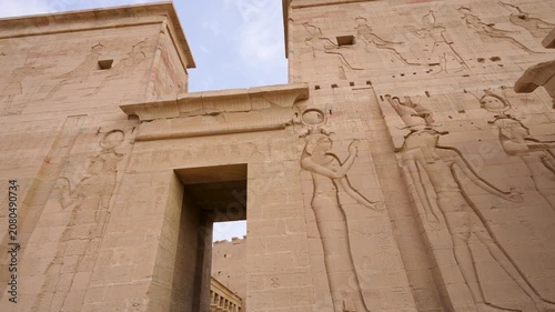 A low angle view of the ancient stone pylon entrance at the Temple of Philae in Aswan, Egypt, featuring intricate hieroglyphic carvings and goddess reliefs.
