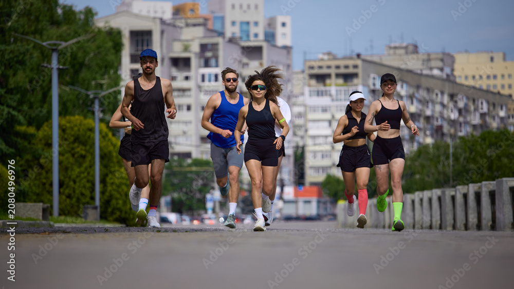 Fototapeta premium Running club jogging toward camera on urban road with city buildings behind. Concept of city fitness, cardio training, endurance, active lifestyle and group motivation.