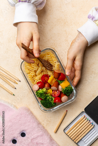 Girl eating Healthy pasta meal prep in glass container with fusilli, sausage, broccoli, cherry tomatoes and bell pepper on table with school supplies, girl hands. Lunch for children, back to school 