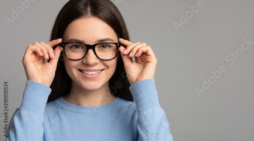Young woman with long dark hair adjusts black eyeglasses while smiling, wearing a light blue sweater against a neutral background