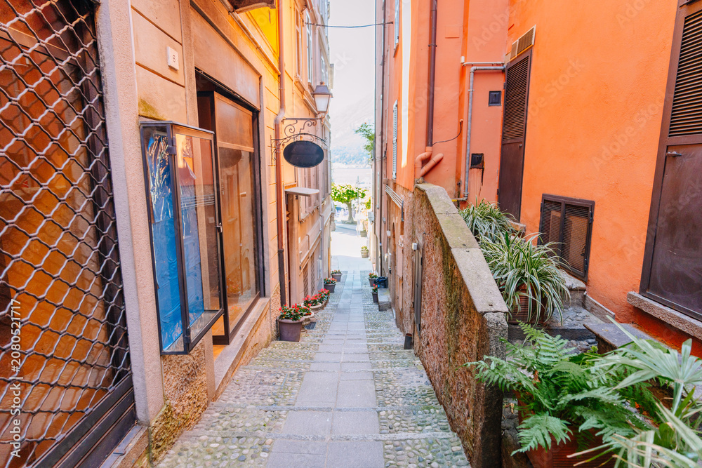 Fototapeta premium Narrow cobblestone alleyway in Bellagio, Italy, lined with colorful buildings and potted plants leading to a scenic view