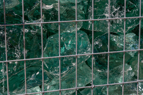 Green glass rocks texture contained inside a metal wire cage gabion wall under bright direct sunlight close up