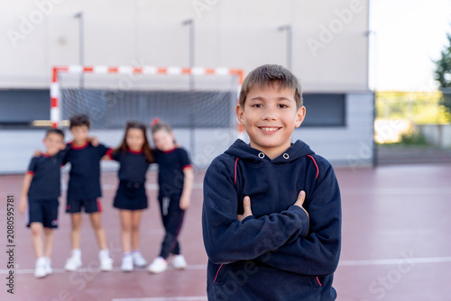 Happy schoolboy standing with arms crossed, classmates blurring in background on a school sports court with a goal