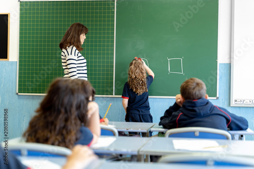 Teacher watching a young female student drawing a circle and square on a green chalkboard during a lesson in a classroom