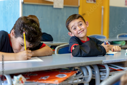 Young male student smiling at desk while writing, engaging in education and childhood development in a school environment