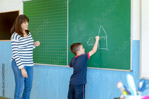 Young boy learning by drawing a house with chalk on a classroom chalkboard while a female teacher watches