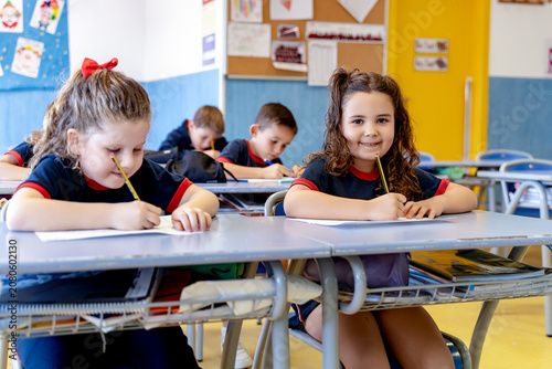 Happy primary students wearing uniform, sitting at desks, writing on paper and studying during school lesson