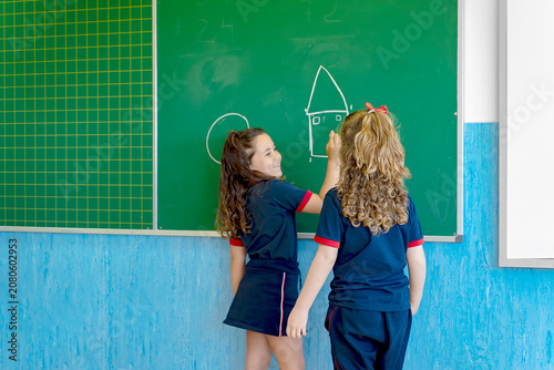 Schoolgirls with school uniforms happily drawing a house and circle on a green chalkboard, creating art in a classroom