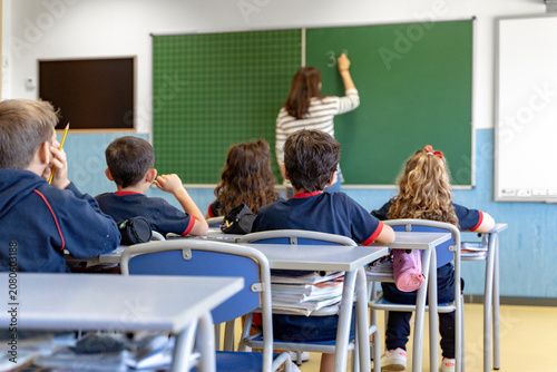 Students sitting at desks in a classroom, listening to a female teacher writing on a green chalkboard during a lesson