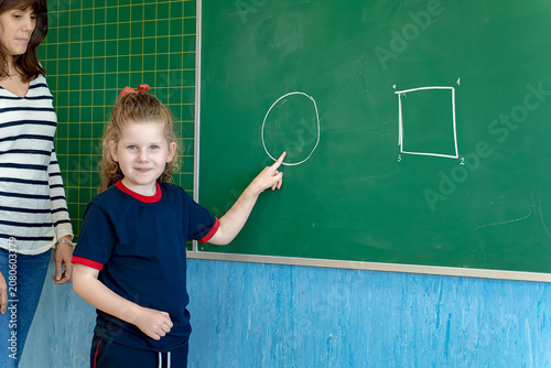 Young female student pointing at a circle drawn on a green chalkboard, a teacher standing nearby supervising her