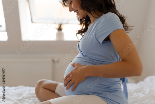 Happy pregnant woman sitting on bed gently touching her baby bump and smiling while enjoying peaceful moments of pregnancy at home, side view