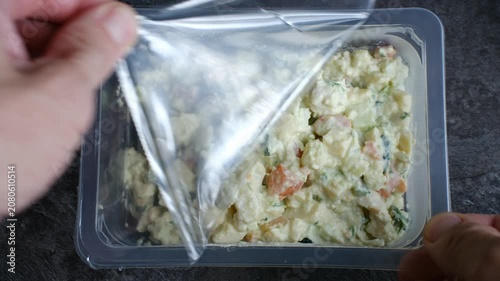 Person hands carefully peeling back plastic film from rectangular container filled with fresh, ready to eat potato salad on dark, textured surface, preparing convenience meal. Semi finished products